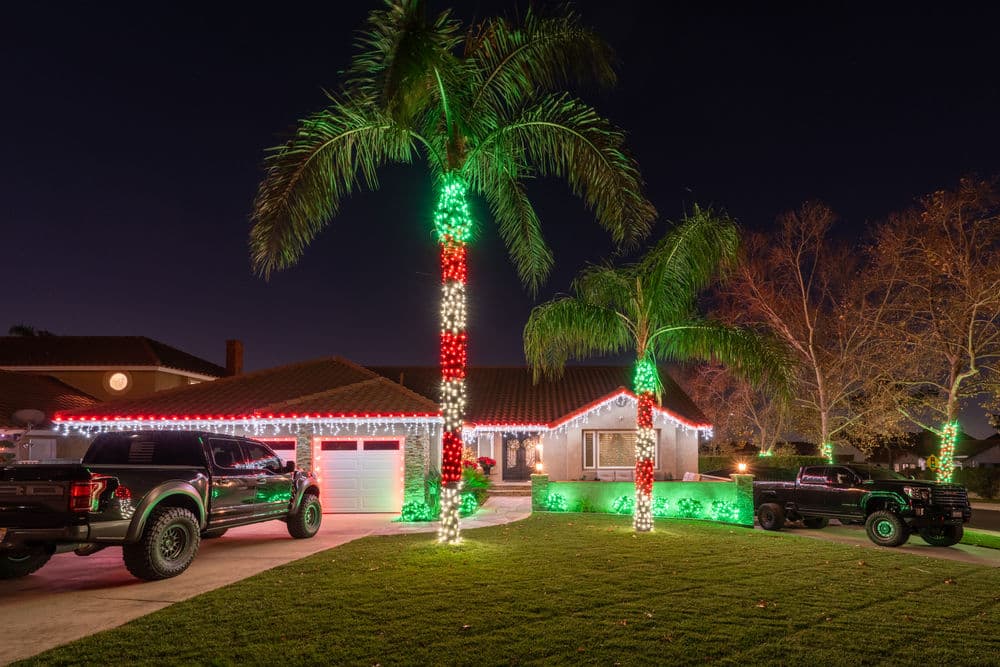 Festively decorated home with vibrant red and green lights, surrounded by palm trees and trucks.