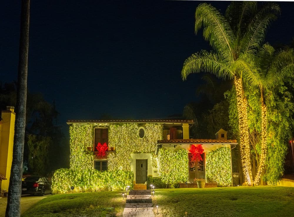 Festively decorated house with lights and large red bows, surrounded by palm trees at night.
